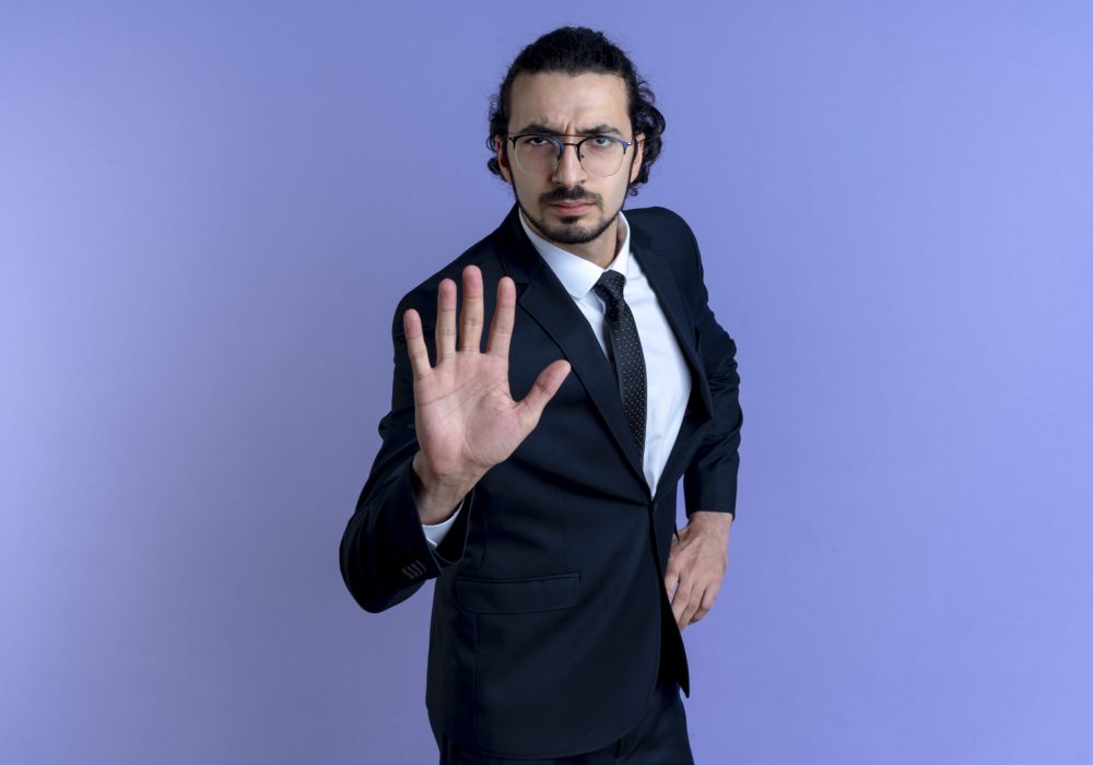 business man in black suit and glasses making stop sign with hand looking at camera with serious face standing over blue background