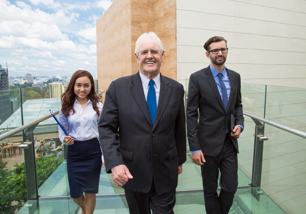 Portrait of happy senior business leader with his male and female assistants walking on open glass bridge between office buildings. Business team concept