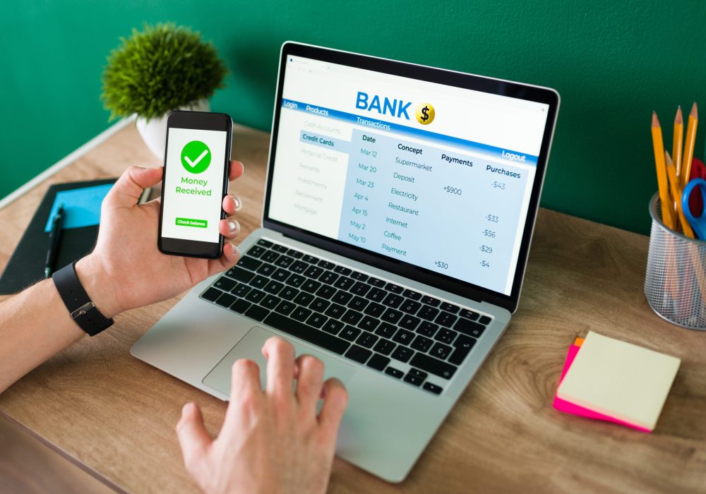 Wire transfer. Young man using his smartphone and laptop to do a banking transaction and some payments