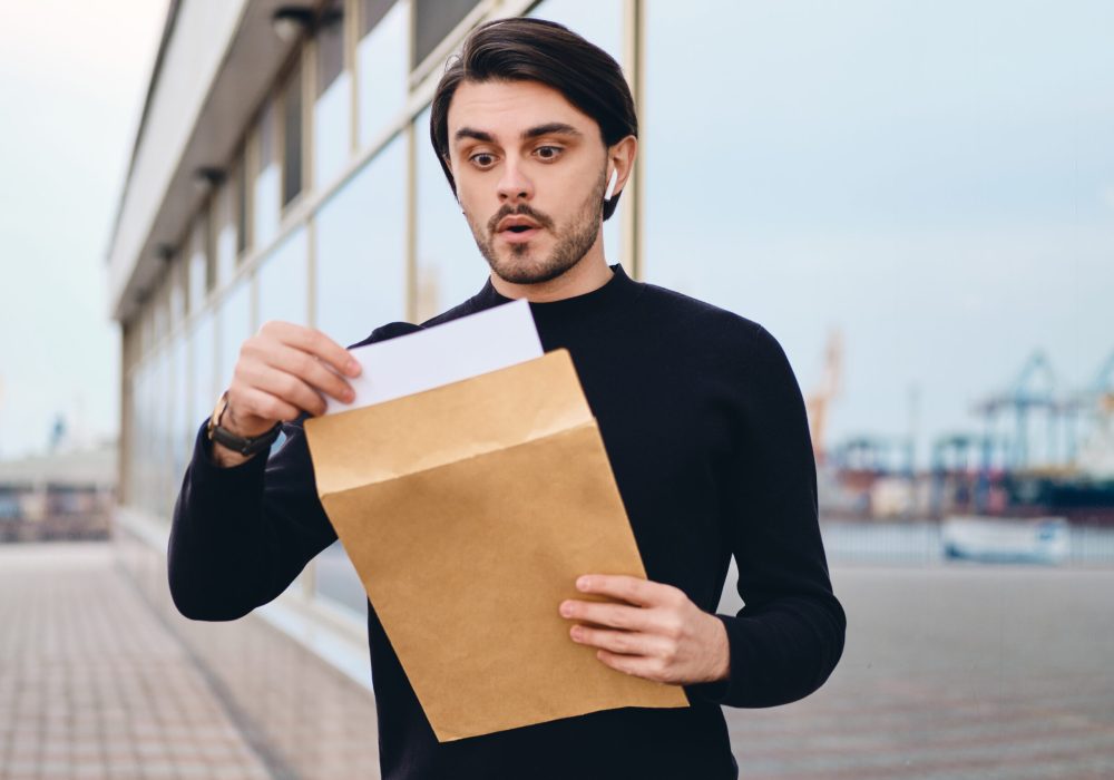 Young excited brunette man amazedly opening envelope with response outdoor