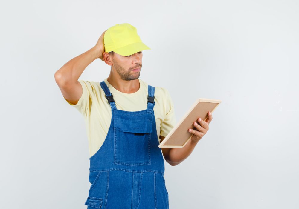 Young loader holding board with hand on head in uniform and looking pensive , front view.